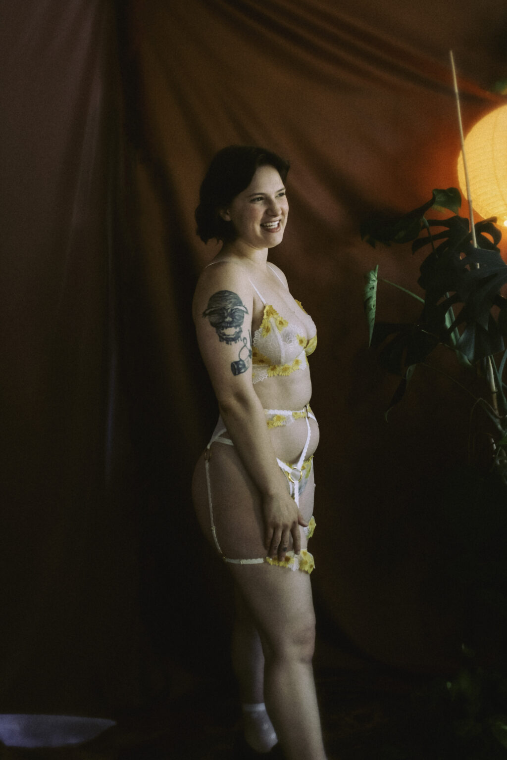 A full body photo of Ivy Maxwell. Ivy is against a dark background, light shining on her as she smiles while wearing a three piece lingerie set.